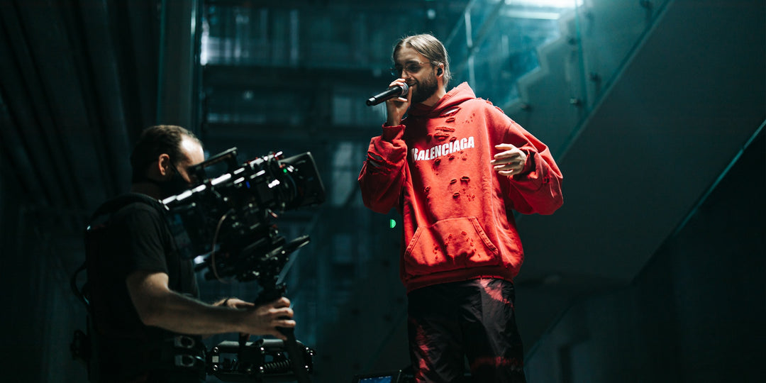 Kevin, gekleed in een rode Balenciaga-hoodie, tijdens een opname met een professionele cameraman. Deze scene benadrukt Kevin's creatieve carrière, waar de inspiratie voor de Mascotte Slowflu KS Vloei + Tips Limited Edition vandaan komt. Het product combineert een urban stijl met topkwaliteit en is een eerbetoon aan Kevin’s relaxte en succesvolle flow, perfect voor liefhebbers van premium rookaccessoires